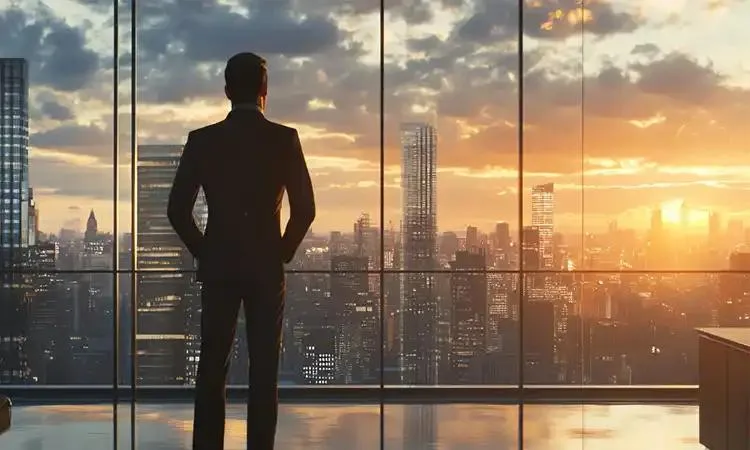 Man in a suit standing with his back to the camera, looking out a large window at a city skyline at sunset.