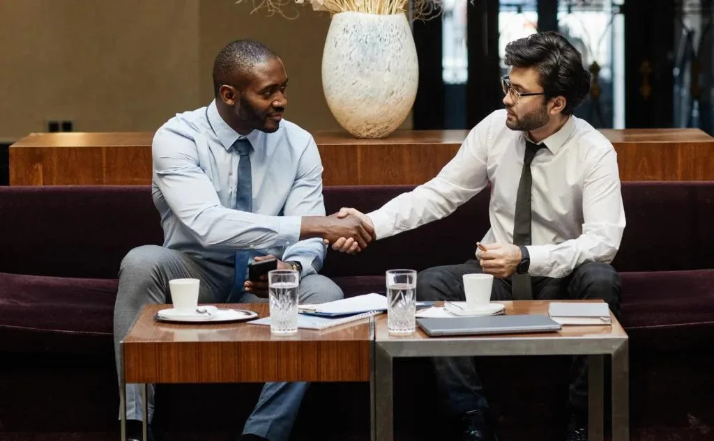Two businessmen in shirts and ties shaking hands over a coffee table in a hotel lounge.