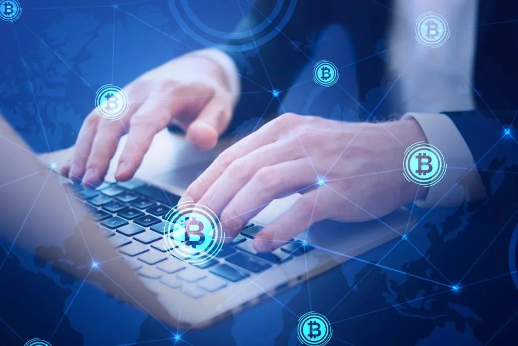 A close-up shot of hands typing on a laptop keyboard, overlaid with a blue digital network grid, world map, and Bitcoin symbols. The image represents blockchain or cryptocurrency technology.