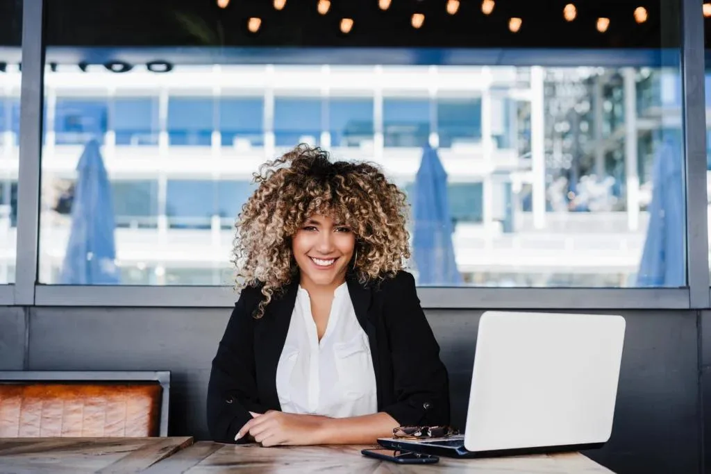 A smiling businesswoman with curly blonde hair is seated at a wooden table with an open white laptop. She wears a black blazer and white shirt. The background is a large window showing a modern outdoor cafe or building.