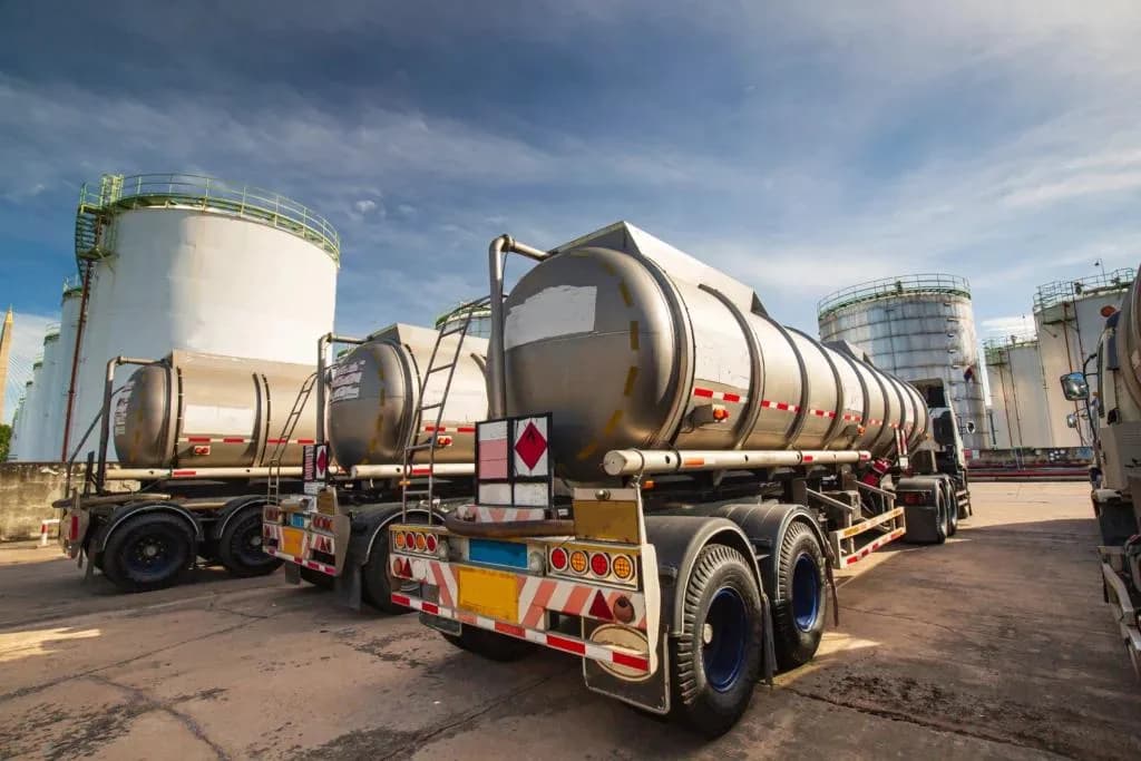 A low-angle view of several stainless steel tank trucks parked in a row, with chemical storage tanks towering in the background. The scene is brightly lit under a blue sky with scattered clouds.