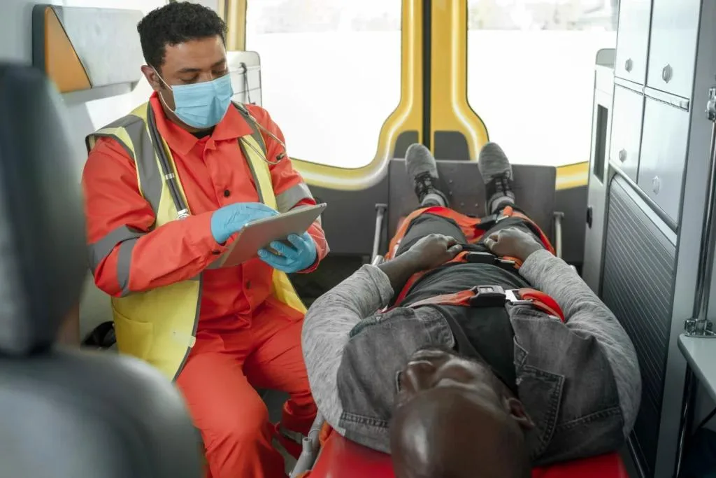 An EMT in an orange uniform, mask, and blue gloves sits in an ambulance, looking at a tablet. A patient is lying on the stretcher, secured with straps.