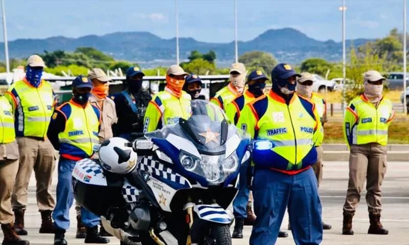A group of South African National Traffic Police officers are standing in formation outdoors. They are wearing high-visibility vests, masks, and uniform caps. A blue and white police motorcycle is parked in the foreground. Mountains are visible in the distance.