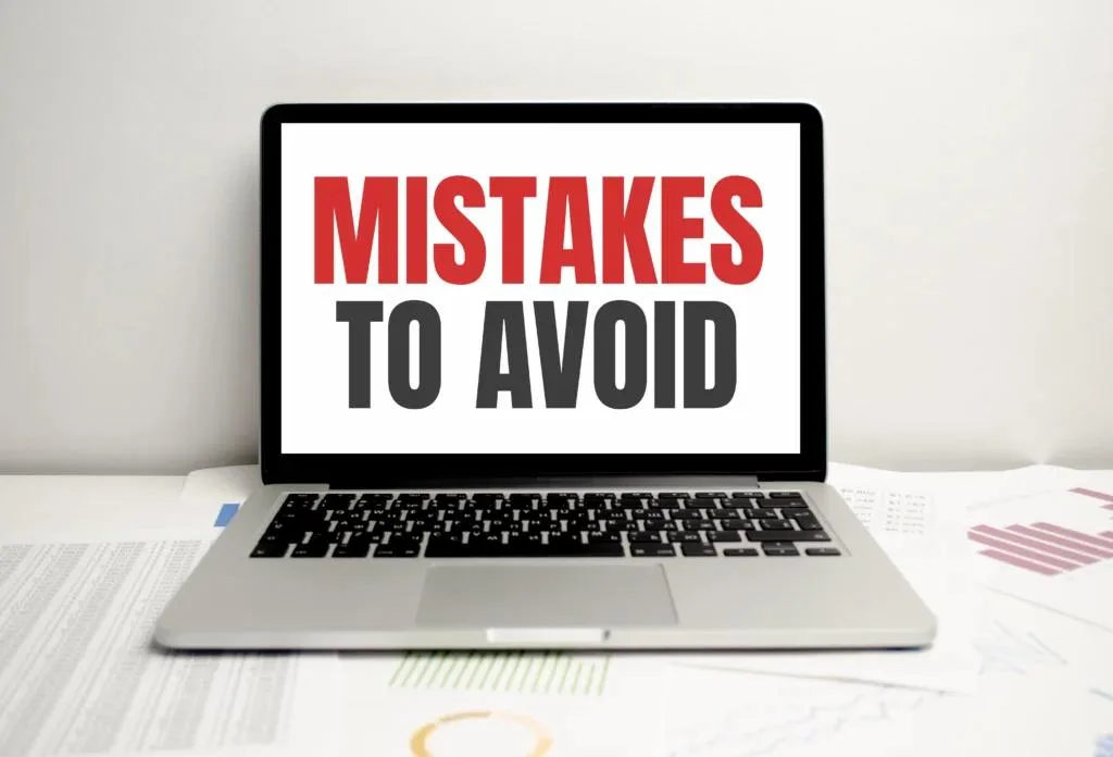 A silver laptop is centered on a desk with paperwork and charts. The screen displays the bold text "MISTAKES TO AVOID" with the word "MISTAKES" in red and "TO AVOID" in dark grey.