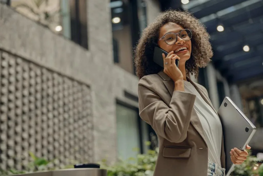 A smiling businesswoman with curly hair and glasses is standing outdoors in a modern urban setting. She's wearing a light blazer, holding a laptop in one hand and talking on a mobile phone with the other.