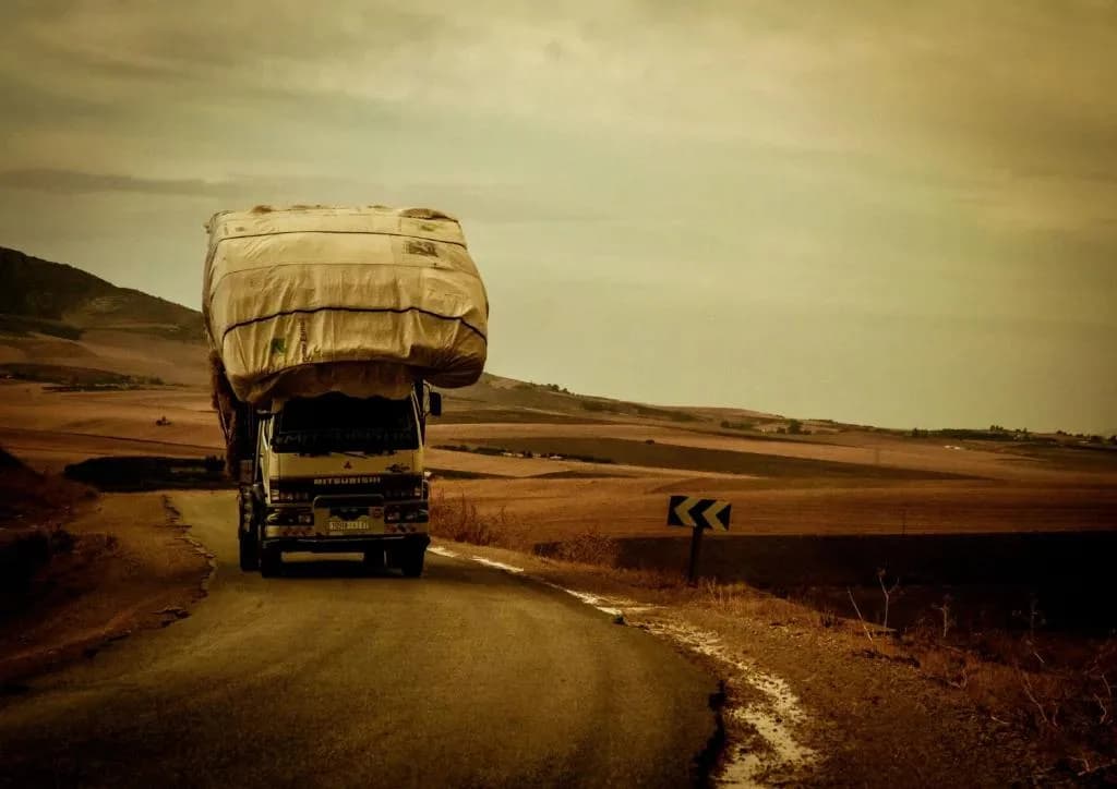 An old Mitsubishi truck is driving down a narrow, dusty road in a dry, rural landscape. The truck is massively overloaded with a large, bulging white sack strapped to the top. The image has a vintage, sepia-toned filter and the sky is a muted yellow-grey. A curved arrow sign is on the side of the road.