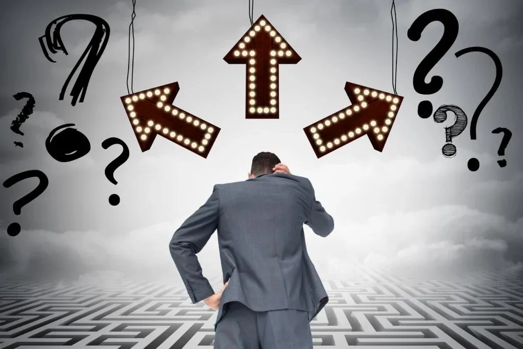 A businessman in a grey suit stands with his back to the camera, scratching his head on a maze-patterned floor. Three lit arrows point left, forward, and right above his head. The background is a cloudy sky with numerous hand-drawn question marks.
