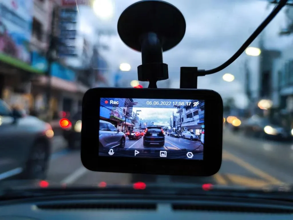 A dash cam is mounted on a car's windshield, displaying a live video feed of a busy street with cars in traffic and buildings. The screen shows the "Rec" status and a date and time stamp. The background outside the windshield is blurry with bokeh lights.