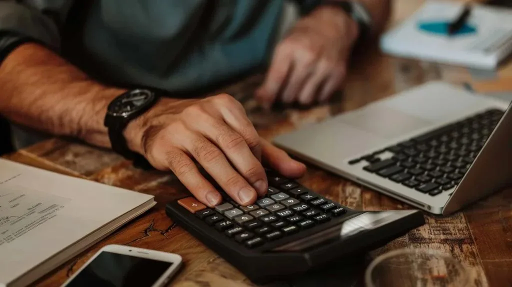 A close-up shot of a person's hands on a calculator, seated at a wooden desk. A wristwatch is on the left wrist, with a laptop and smartphone also on the desk.
